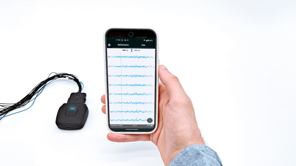 Hand holding a smartphone showing real-time EEG data streaming on the Mentalab Mobile App from the Explore Pro device. In the background, the Mentalab Explore Pro device is connected to electrode cables.